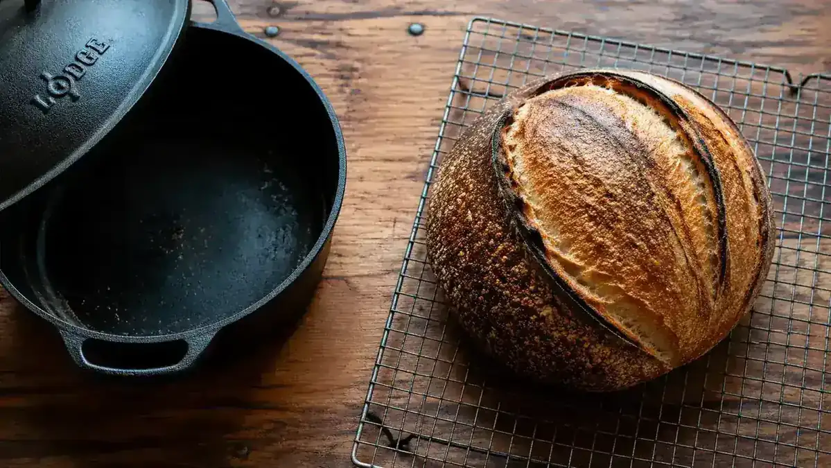 A round sourdough loaf with a pronounced ear and dark caramelized crust cooling on a wire rack next to a Lodge cast iron Dutch oven
