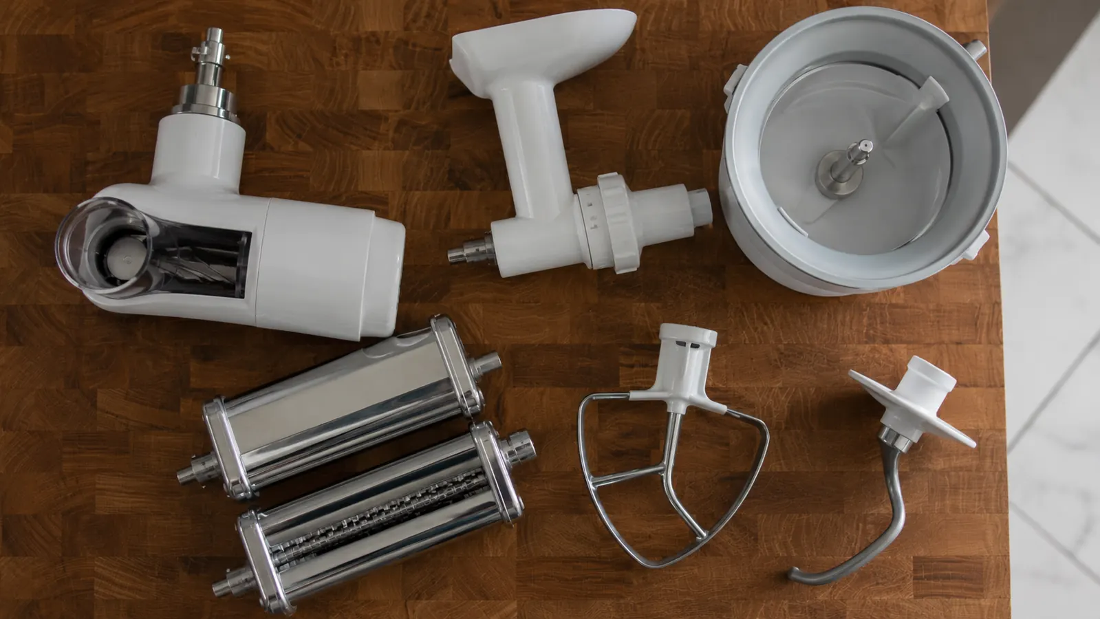 Spread of KitchenAid attachments on a kitchen counter including pasta roller, grain mill, and meat grinder with price tags suggesting the total cost